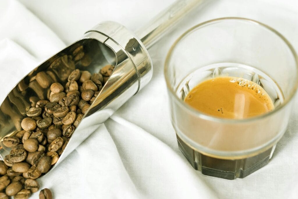 a clear drinking glass with coffee near the stainless scoop with coffee beans