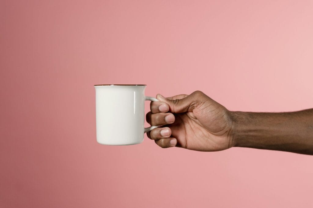 Minimalist studio shot of a hand holding a white mug on a pink backdrop.