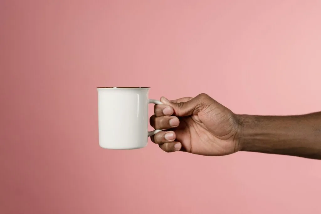 Minimalist studio shot of a hand holding a white mug on a pink backdrop.