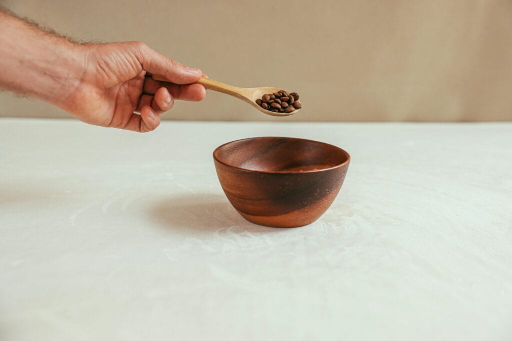 Close-up of a hand holding a spoon with coffee beans over a wooden bowl on a white table.