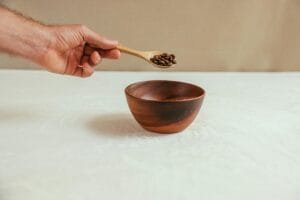 Close-up of a hand holding a spoon with coffee beans over a wooden bowl on a white table.