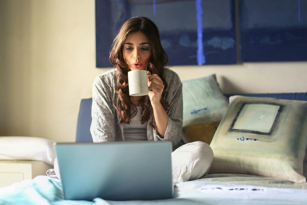 A woman enjoying her morning coffee while working from home in a cozy bedroom setting with a laptop.